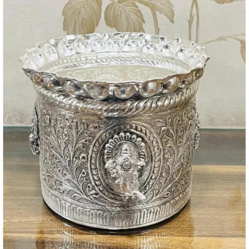 An intricately designed silver decorative container with ornate floral patterns and embossed religious figures, placed on a wooden surface. The container features a detailed lid with a scalloped edge, showcasing traditional craftsmanship.