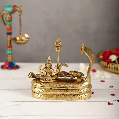 Brass sculpture depicting a deity reclining on a serpent, with a multi-headed hood, set on a decorative platform with a traditional oil lamp in the background.