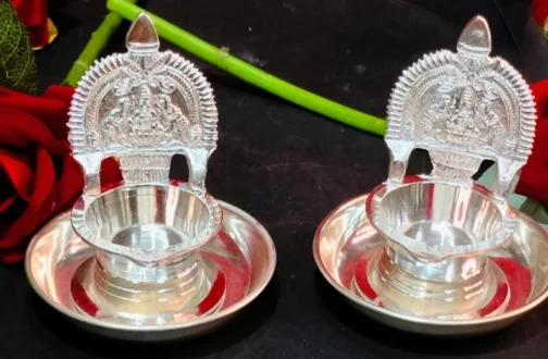 Two intricately designed silver decorative bowls with deity-engraved backrests, placed on a circular base. The setup includes red roses and green stems in the background, creating an elegant display.