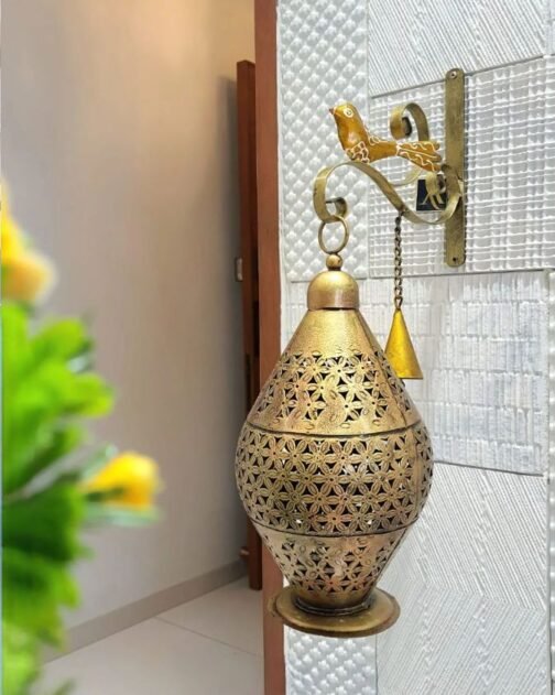 Decorative golden metal wall lantern with intricate floral cutouts, hanging from a wall-mounted bracket featuring a small bird design and a hanging bell. The lantern is set against a textured white wall, adding a touch of elegance to the space.