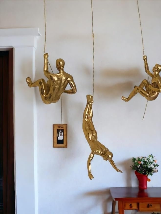 Three gold figurines hanging from ropes, styled as climbers, creating a dynamic wall decor piece. The installation is set against a white wall, with a small red table holding a vase of flowers underneath, adding a modern and artistic flair to the space.