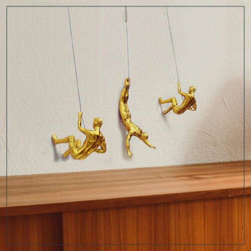 Three gold climber figurines hanging on ropes against a white wall, with each figure posed in a climbing action. The arrangement creates a lively and artistic decor piece, adding a unique and modern touch above a wooden console.