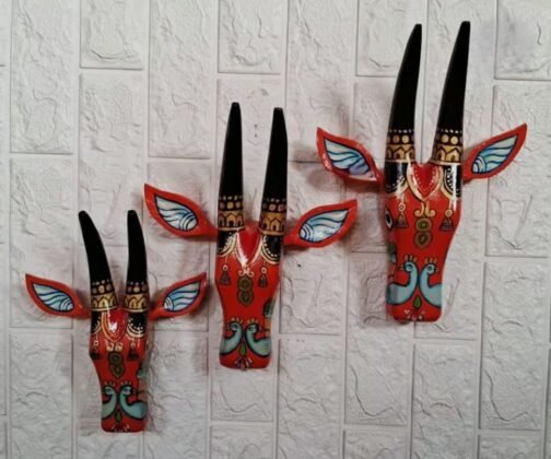 Three hand-painted wall masks with red bases, featuring tall black horns, blue and red accented ears, and intricate traditional patterns. Each mask showcases a unique design with stylized bird motifs, adding a bold and cultural touch to the wall.