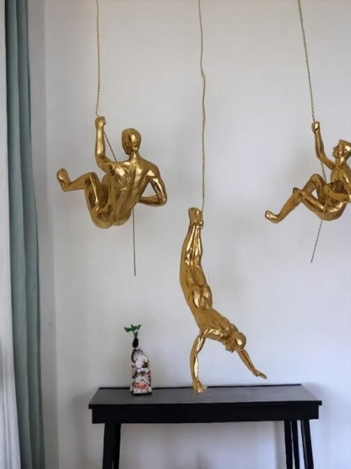 Three gold climber figurines hanging on ropes in different poses, creating an artistic wall display above a black console table. A small decorative sculpture is placed on the table, adding an additional element of art and style to the minimalist setup.