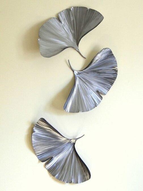 Three silver leaf-shaped wall art pieces arranged vertically on a light-colored wall. The metallic leaves have a ribbed texture and a fluid, fan-like design, creating an elegant and contemporary look that enhances any interior space.