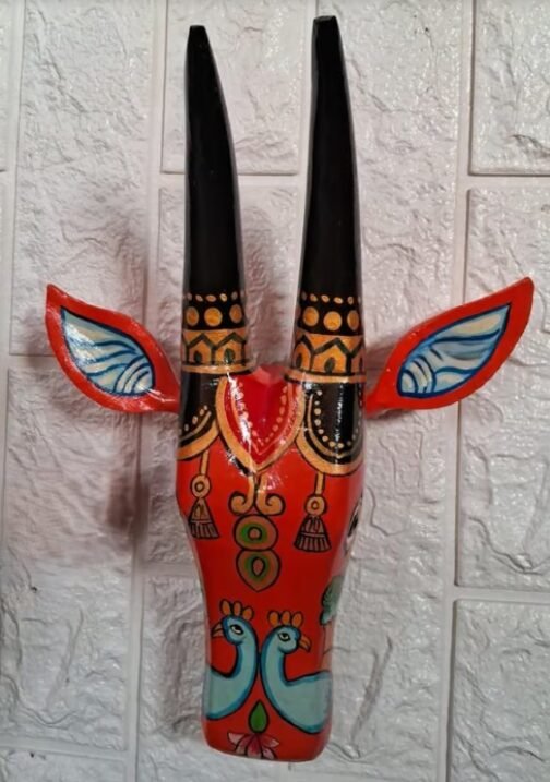 A close-up of a red hand-painted wall mask featuring two tall black horns with gold dot patterns, blue and red accented ears, and intricate designs with stylized birds and floral motifs. The mask is mounted on a textured white wall, showcasing detailed traditional artistry.