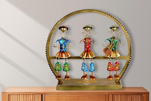 A decorative metal art piece featuring three colorful tribal figurines with intricate detailing, dressed in blue, red, and green outfits. The figurines are placed within a circular golden frame, positioned on a wooden surface, adding a vibrant touch to the room decor.