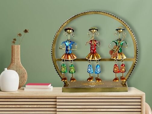 Decorative metal sculpture featuring three tribal figurines in vibrant blue, red, and green outfits, set within a circular golden frame on a wooden console. The scene is complemented by a wicker vase with dried flowers and a stack of books, creating an artistic and cultural decor display.