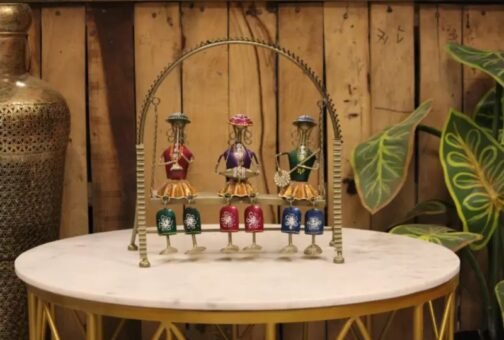 A metal sculpture showcasing three colorful tribal figurines in red, purple, and green attire, set within a golden arch on a round marble table. The decor is complemented by a large decorative vase and leafy green plants in a rustic setting, adding a traditional and artistic touch to the space.