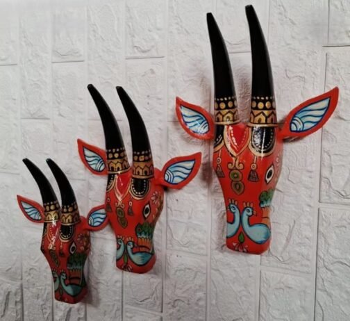 Three red hand-painted wall masks with tall black horns, blue and red accented ears, and intricate traditional patterns featuring stylized birds and floral designs. The masks are mounted on a textured white wall, adding a bold and cultural element to the decor.